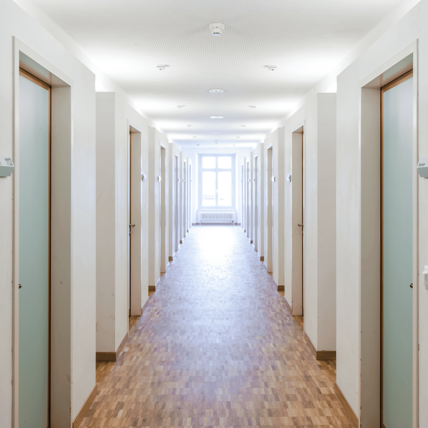 Bright, empty corridor with white walls, wooden floor, and multiple doors leading to a window.
